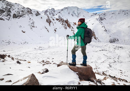 Hiker with trekking poles and backpack standing on the rock at snowy mountains background Stock Photo