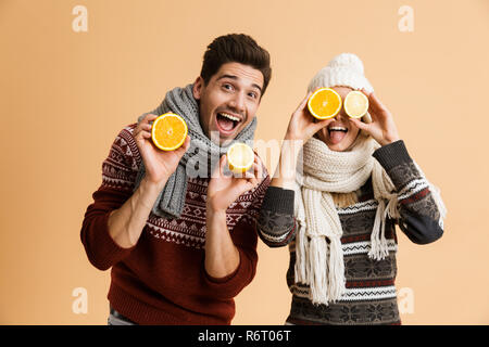 Portrait of a smiling young couple dressed in sweaters and scarves standing together isolated over beige background, showing sliced lemons and oranges Stock Photo