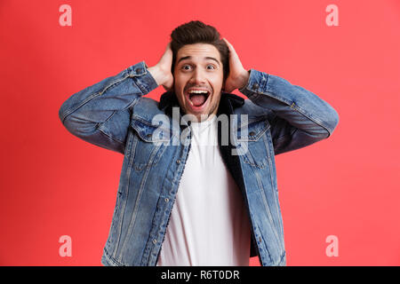 Portrait of an excited young man dressed in denim jacket standing isolated over red background, screaming Stock Photo
