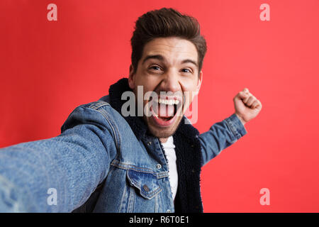 Portrait of a cheerful young man dressed in denim jacket standing isolated over red background, taking a selfie Stock Photo