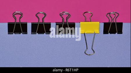 A side view of a line of black binder clips with a single yellow one to break up the conformity. Clips are attached to a purple folder. Stock Photo