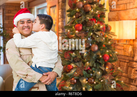 Hispanic Armed Forces Soldier Wearing Santa Hat Hugging Son In Front Christmas Tree. Stock Photo