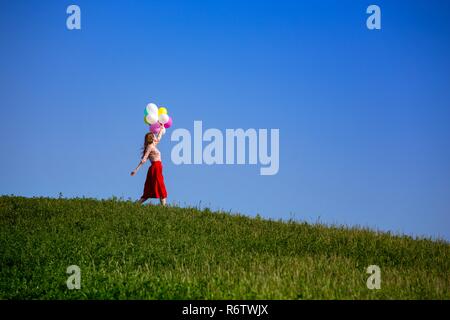 Happy girl in the meadows tuscan with colorful balloons, against the blue sky and green meadow. Tuscany, Italy Stock Photo