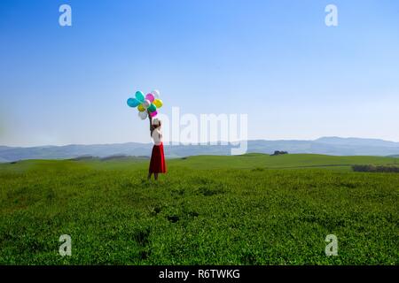 Happy girl in the meadows tuscan with colorful balloons, against the blue sky and green meadow. Tuscany, Italy Stock Photo