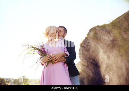 Couple in love stands in the splashes of water against the sunset. A man hugs a woman in a beautiful dress. Love in the eyes Stock Photo
