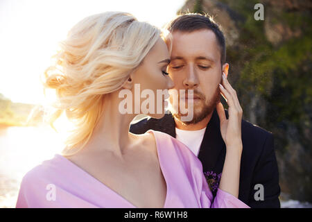 Couple in love stands in the splashes of water against the sunset. A man hugs a woman in a beautiful dress. Love in the eyes Stock Photo