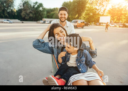 Young dad carries mom and son in a cart on the parking lot Stock Photo