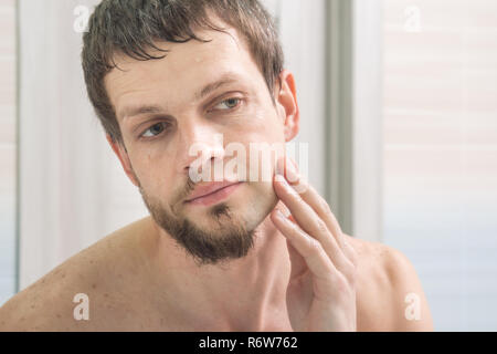 The guy shaved one half of his face and looks in the mirror evaluating the result Stock Photo