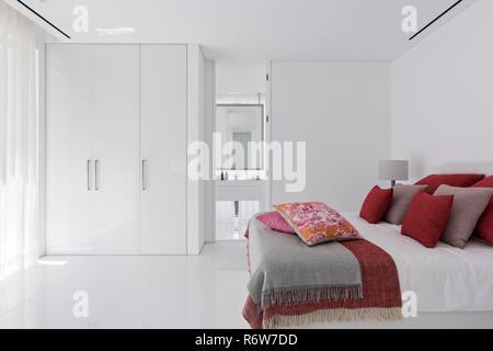 Red and muted pink cushions on double bed with built-in wardrobe in minimalist bedroom of  new build villa, Quinta do Lago Stock Photo