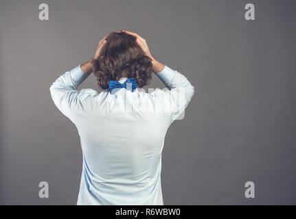 Backside view standing young man with shaggy curly hair wearing blue shirt and bow tie against grey background. Holding head, depression. Negative despair emotion concept. Rear view people collection. Stock Photo