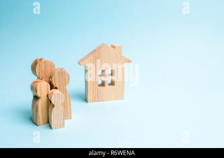 family stands near a wooden house on a blue background. The concept of affordable housing and mortgages for buying a home for young families and coupl Stock Photo