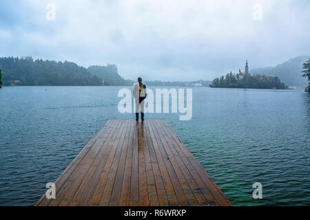 traveler man standing on the lake wooden pier in fog against island with church. travel lifestyle concept. Bled lake, Slovenia Stock Photo