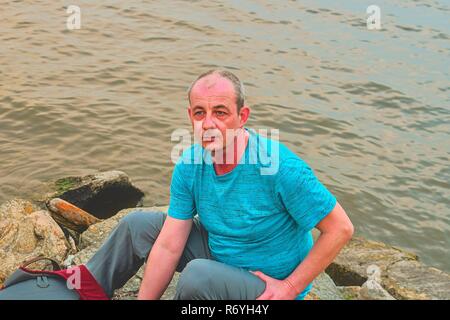 Midle aged man sitting on the shore of the lake. Solitude mature man sitting on the bank. Concept of loneliness