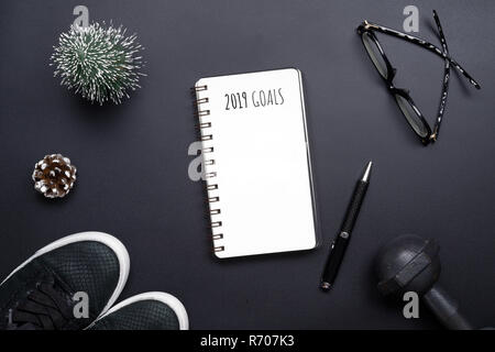 2019 new years resolutions sport flat lay composition. Black shoes, black dumbbells, glasses, blank notebook and Christmas decoration on black backgro Stock Photo