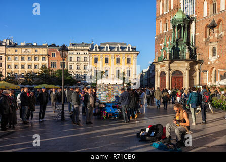 City of Krakow in Poland, people and busker on lively Main Market Square in the Old Town in front of St. Mary Church, sunset time, Stock Photo