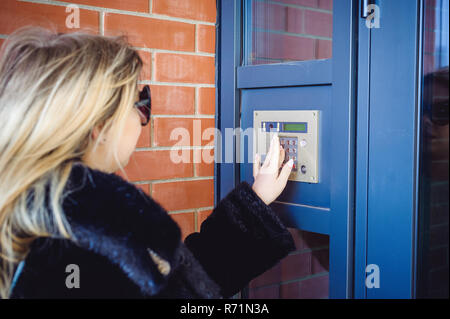 young blond woman in a fur coat, dials the code on the doorphone panel, presses  buttons with her fingers to get inside house Stock Photo