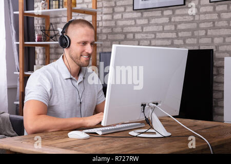 Smiling Young Man Using Headphone While Working On Computer Over Wooden Desk Stock Photo