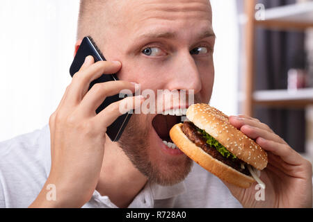 Portrait Of A Young Man Talking On Mobile Phone While Eating Burger Stock Photo