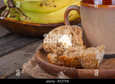 Closeup of a banana nut muffin cut in half with a coffee cup on a wooden plate and bowl of bananas in background Stock Photo