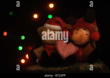 Little Christmas teddy bear and reindeer sitting on moss and holding a heart, in front of Christmas lights. Stock Photo