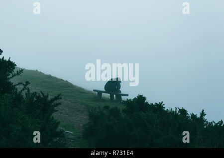 A sad hooded man sitting on a bench looking away and down from the camera on a misty, moody day Stock Photo
