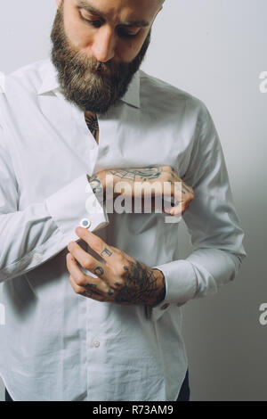 Bearded young man wearing white shirt, doing up cuff links, tattoos on hands Stock Photo