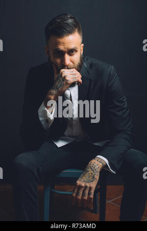 Portrait of young man, sitting on chair, wearing suit, tattoos on hands, pensive expression Stock Photo