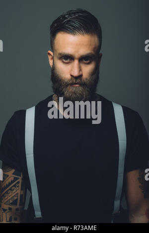Portrait of young man with beard, wearing trouser braces, tattoos on arms Stock Photo