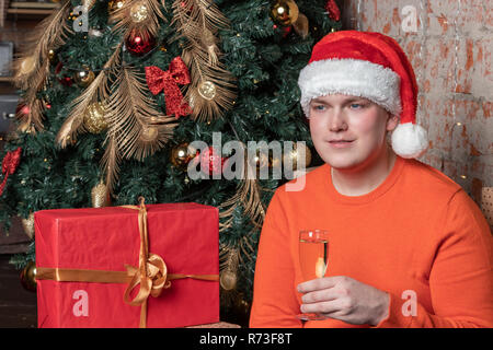 Handsome guy in santa claus hat holds a glass of champagne sitting under the tree surrounded by boxes of gifts Stock Photo