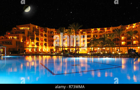 Moon above resort hotel and swimming pool at night. Pool and evening hotel on vacation. Lights of evening hotel are reflected in pool water in night.  Stock Photo