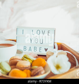 Close up Romantic Breakfast in bed with I love you baby text on lighted box. Cup of coffee, juice, macaroons, flower and gift box on wooden tray. Birt Stock Photo