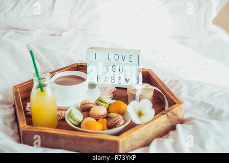 Romantic Breakfast in bed with I love you baby text on lighted box. Cup of coffee, juice, macaroons, flower and gift box on wooden tray. Birthday, Val Stock Photo