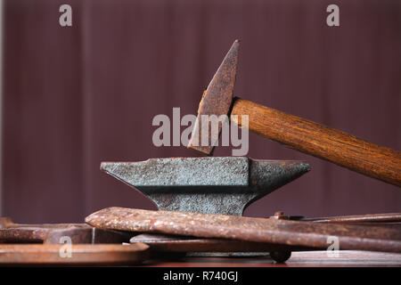 Old rusty rugged anvil, hammer and other blacksmith tools on brown natural wooden background. Stock Photo