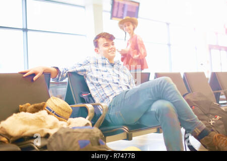 Young man sitting at airport, backpack beside him, young woman standing nearby, looking towards him Stock Photo
