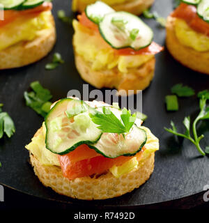 Close up of sandwiches with salmon, scrambled eggs and cucumber slices on black wooden board. Stock Photo