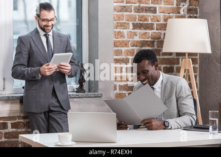 handsome businessman standing by window while african american partner sitting at table and reading document Stock Photo