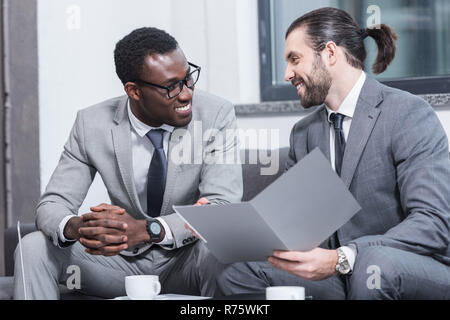 smiling multiethnic business partners sitting on couch and talking in office Stock Photo