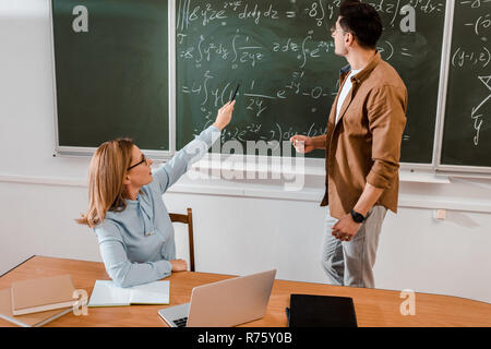 Female teacher pointing at equations while student standing near chalkboard Stock Photo