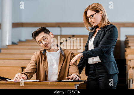 female university teacher looking at male student in glasses writing exam in classroom Stock Photo