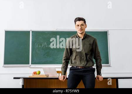 serious male teacher in formal wear looking at camera and standing near desk in classroom Stock Photo