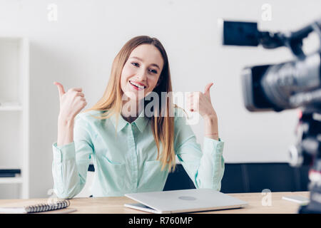 smiling attractive female video blogger recording vlog and showing thumbs up in office Stock Photo