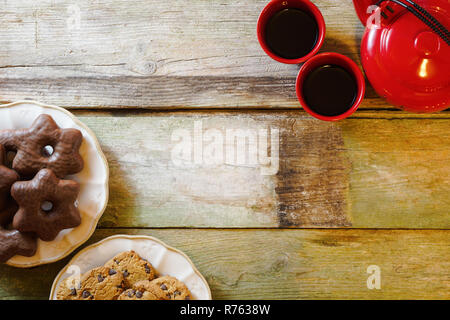 Flat lay of rustic wooden table with chocolate chip cookies in ceramic plates, red teapot and cups. With copy space. Stock Photo
