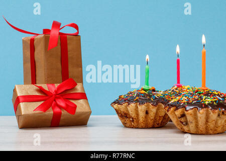Gift boxes and three tasty birthday cupcakes with chocolate glaze and caramel, decorated with burning festive candles on blue background. Happy birthd Stock Photo