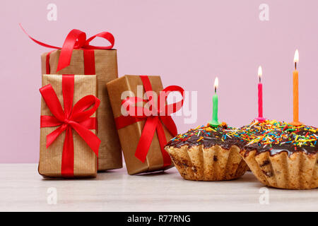 Gift boxes and three tasty birthday cupcakes with chocolate glaze and caramel, decorated with burning festive candles on pink background. Happy birthd Stock Photo