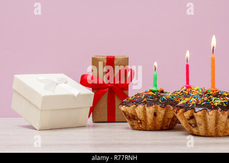 Gift boxes and three tasty birthday muffins with chocolate glaze and caramel, decorated with burning festive candles on pink background. Happy birthda Stock Photo