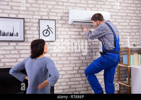 Young Woman Looking At Male Technician Repairing Air Conditioner Mounted On Brick Wall Stock Photo