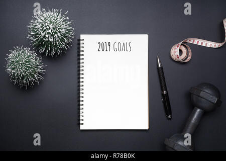 2019 new years resolutions sport flat lay composition. Black dumbbells, measurement tape, blank notebook and mini Christmas tree on black background.  Stock Photo