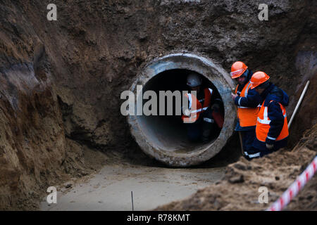 three workers install concrete gutters on the side of the road, power plants. Frame lifting concrete drainage pipe. Russia Berezniki 23 November 2017 Stock Photo