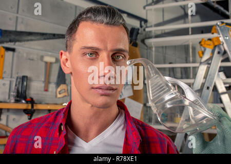 young handsome caucasian carpenter holds safety glasses in his hands in his workshop Stock Photo