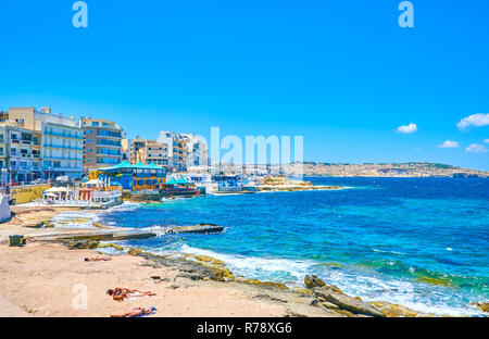 BUGIBBA, MALTA - JUNE 14, 2018: The small sand beach among rocks in the center of resort is a popular place to have a sunbaths, on June 14 in Bugibba. Stock Photo